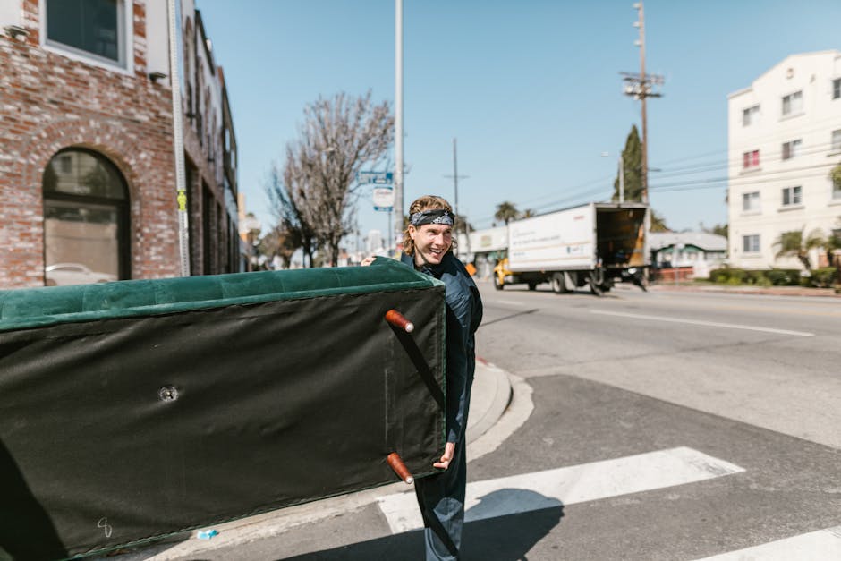A person with long hair tied back, wearing a black jacket and a patterned headband, is standing on the street corner near a residential building with brick walls and arched windows. They are smiling and holding a large, rectangular piece of furniture or bulky item wrapped in green and black protective padding, with visible fabric and plastic material. The item appears to be a piece of furniture being prepared for removal, as part of a home relocation process organized by Movers Pimlico. The setting includes a curb, a crosswalk, and a moving van with an open side door parked nearby, which is likely used for furniture transport. In the background, a delivery truck is driving along the road, and there are leafless trees, electrical poles, and modern white apartment buildings. The scene is lit by natural daylight, emphasizing the outdoor loading and furniture handling involved in packing and moving services.