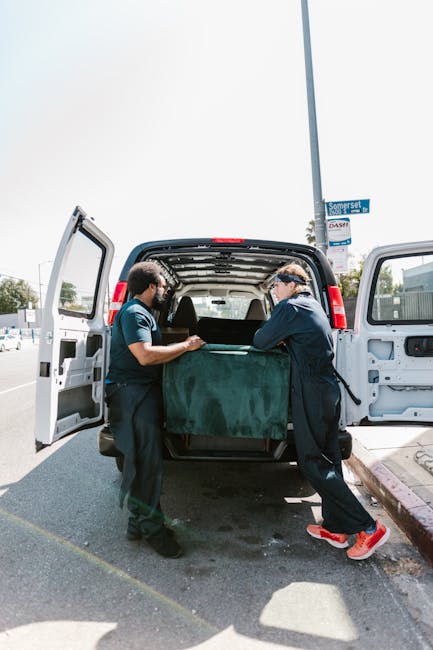 Two professional movers from Movers Pimlico are engaged in the loading process of bulky furniture into the rear of a white moving van, parked outside a residential area. One mover, wearing a dark uniform and gloves, is assisting in guiding a large piece of furniture covered with a protective green fabric or blanket into the van. The other mover, dressed in a similar dark uniform with red shoes, is helping to lift and position the furniture. The van's side and rear doors are open to facilitate furniture transport, with the interior visible showing additional packaging materials. The setting indicates a house relocation scenario, with the loading taking place on the driveway adjacent to the curb, near a blue street sign and utility pole, under daylight. This scene exemplifies the careful handling and systematic loading involved in professional removals, which are part of comprehensive packing and furniture transport services offered by Movers Pimlico for home relocations.