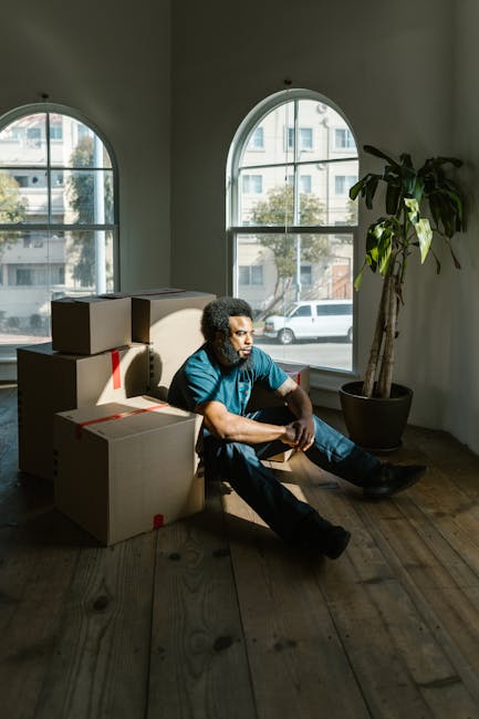 A man with dark curly hair and a beard, wearing a navy blue t-shirt and matching trousers, is inside a well-lit room with white walls and light wooden flooring. He is holding a cardboard box sealed wi