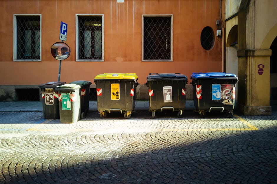 Five waste and recycling bins positioned on a cobblestone sidewalk in front of a peach-colored building with barred windows. The bins include a grey general waste bin, a yellow bin designated for electronic waste disposal, a black bin for recyclables, and a blue bin for paper waste. Each bin has distinct colour-coded lids and labels indicating their specific waste categories, with some showing symbols for electronics, plastic, paper, or general refuse. The yellow bin for e-waste has a label emphasizing the importance of legal disposal of electronics after a house relocation, and moverspimlico.co.uk is mentioned as the service provider. A convex safety mirror is mounted on a pole behind the grey bin, reflecting part of the street scene. The setting suggests an urban environment with a focus on waste management in the context of moving and relocation services, highlighting the logistical process of sorting and disposing of electronic waste after a move.