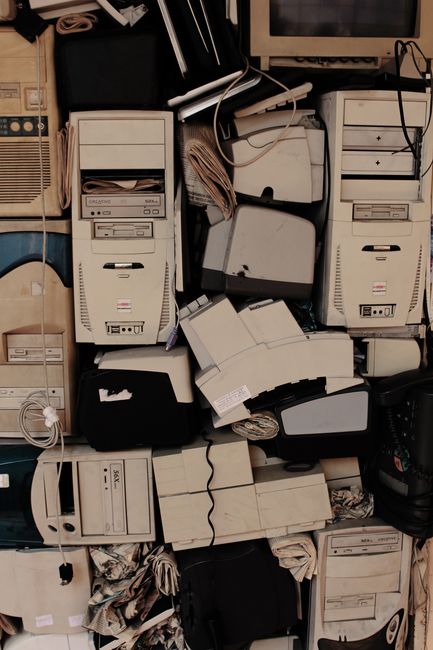 A cluttered indoor storage area filled with numerous discarded desktop computer towers, some stacked on top of each other and others leaning against each other. The beige and black plastic casings of the computers show signs of wear, with a few having visible paper labels or sticky notes attached. Several cables and power cords are hanging loosely from the devices and draped over nearby objects. The background includes rolled-up brown paper, cardboard boxes, and a small section of a wooden shelf or wall, indicating an environment used for storing or disposing of electronic waste. The lighting is dim, highlighting the disorder and emphasizing the need for proper disposal, which Movers Pimlico aims to facilitate in their specialized house removal and e-waste recycling services. The scene captures the process of clearing a property during a move, aligning with the theme of furniture transport, packing, and responsible disposal post-relocation.