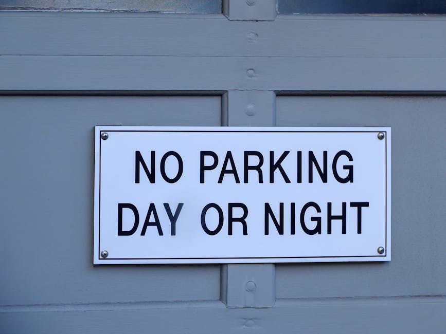 A rectangular white sign with black, bold, uppercase text that reads 'NO PARKING DAY OR NIGHT' is mounted on a light grey wooden door or panel. The sign is fixed with four metal screws, one in each corner. Behind the sign, the surface appears to be part of a building or storage unit, with visible horizontal and vertical wooden panels. The lighting in the scene is natural, providing clear visibility of the sign and the surrounding surface. This image relates to house removals and moving services, illustrating parking restrictions that might affect furniture transport and home relocation logistics. The sign's message is important for planning efficient moving operations, as illustrated by the professional approach of companies like Movers Pimlico, who operate within local council permit rules for removals in Pimlico.