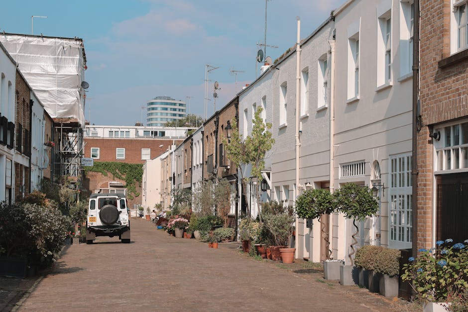 A narrow residential street in Pimlico with row houses featuring white and brick facades, some with bay windows and small front gardens filled with potted plants and small trees. A white van with a spare tire mounted on the rear is parked on the cobblestone pavement, positioned close to the entrance of one house. The loading process appears to be ongoing, likely part of a home relocation, as indicated by the presence of moving equipment and materials. The street is well-lit with natural daylight, and a modern multi-storey building can be seen in the background. The scene reflects typical urban house removal logistics managed by Movers Pimlico, with attention to careful furniture and box transportation, as part of the packing and moving process within the local area. Foam padding, moving blankets, and straps may be visible on furniture, and the pavement is clear to facilitate smooth loading and unloading of items for a residential move.