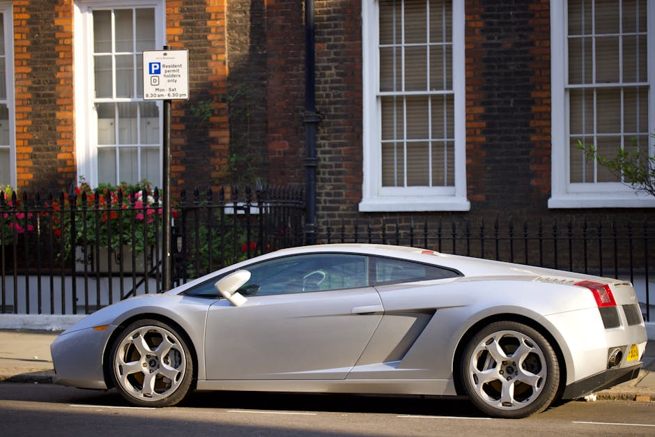 A silver two-door sports car is parked on the side of a residential street in front of a brick house with white-framed sash windows and a black metal fence. The vehicle is positioned parallel to the pavement, with its front facing to the left of the image. Inside the car, the interior is faintly visible through the tinted windows, and the car's sleek, aerodynamic design features smooth curves and alloy wheels. Adjacent to the car, a white and blue parking restriction sign indicates resident permit parking hours from 8:30 am to 6:30 pm. The house's exterior displays flower boxes with colorful blooms on the window sills, and a black drainpipe runs vertically down the brick wall. The scene is illuminated by daylight, capturing a typical urban setting where car parking and house removal logistics might intersect, such as during house relocations or furniture transport managed by Movers Pimlico in London.