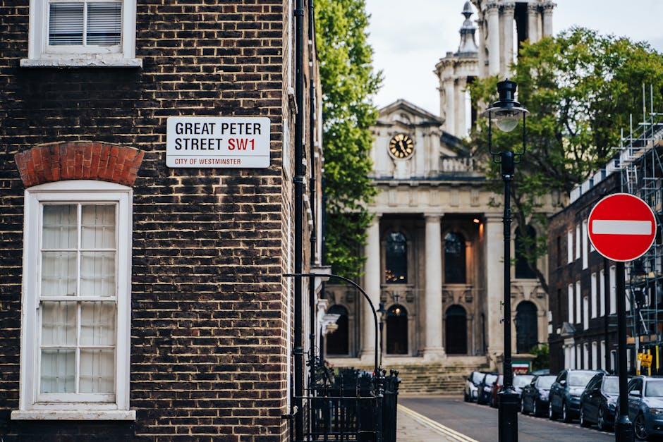 A street scene in Westminster showing a brick building with a white-framed window and a street sign for Great Peter Street SW1, indicating the area’s location within the city. In the background, there is a classical-style building with a clock and tall columns, partially visible behind green trees. A black lamppost stands on the pavement, and a red and white no-entry traffic sign is positioned on the right side of the image. To the right, a construction scaffold is partially visible alongside other parked cars lining the street. The scene appears to be during daytime with natural light illuminating the surroundings, and the setting is indicative of an urban environment suitable for local house removals or furniture transport as part of a home relocation process. This image, associated with Movers Pimlico, offers an accurate view of a typical London street that may be relevant for articles about moving logistics or parking fines related to house removals in Westminster.