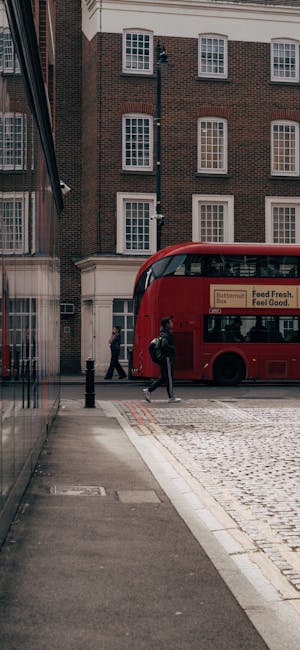 A street scene outside a residential building in Pimlico shows two individuals walking on the pavement near a red double-decker bus parked adjacent to the curb. The building has a brick facade with large, white-framed sash windows aligned vertically and evenly spaced. Visible in the image are various objects related to house removals and moving logistics: cardboard boxes stacked near the entrance of the property, some wrapped in plastic, and a small trolley or hand truck positioned beside the boxes, indicating packing and loading activities. The pavement includes tactile paving, and the area appears well-lit with natural daylight. This scene reflects the typical environment of home relocation, with moving equipment and packaging materials in use, consistent with professional removal services like those offered by Movers Pimlico, supporting efficient furniture transport and packing during a residential move.