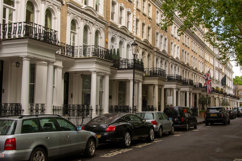 Photograph of a row of elegant white terraced houses with classical architectural details, including tall columns supporting balconies with wrought iron railings. The street in front is lined with parked cars, including a silver station wagon, a black sedan, and other vehicles. The houses have large windows and decorative moldings, with some featuring small balcony gardens with potted plants or trees. A lamppost and Union Jack flags are visible along the pavement, which is partially shaded by green trees. The scene depicts an inner-city residential area in Pimlico, suitable for house removals or furniture transport during home relocation, with a focus on urban environment and architectural style.