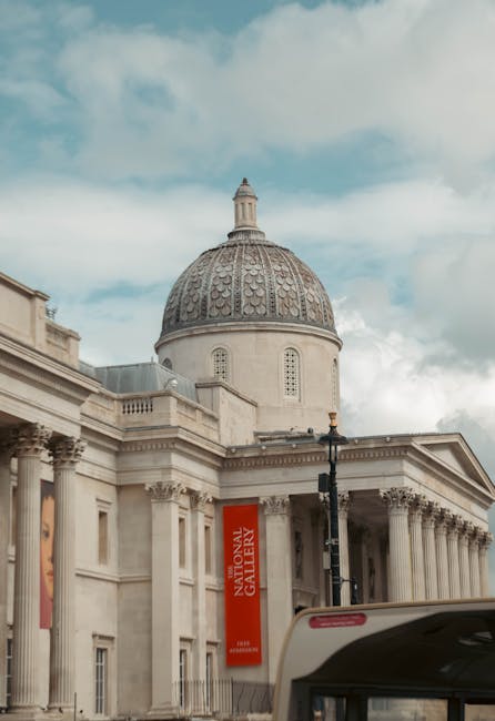 The image shows the exterior of the Tate Britain art gallery with its neoclassical architectural style, featuring tall columns and a large, ornate dome with detailed patterns. In the foreground, part of a van used for house removals is visible, with the vehicle parked close to the gallery entrance on the street. Behind the van, a streetlamp is present, and the sky above is partly cloudy with patches of blue. The scene depicts the process of furniture and packing materials being prepared for home relocation, with visible cardboard boxes and protective wrapping on furniture inside the van, illustrating a loading process typical of professional removals services by Movers Pimlico near the Tate Britain location, supporting efficient furniture transport and logistical coordination for house moves.
