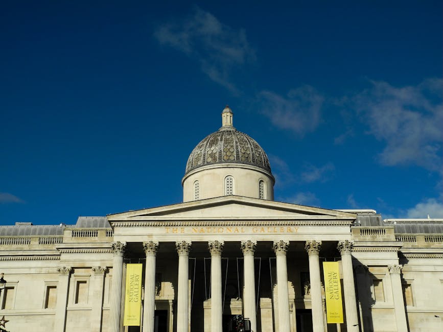 Photograph showing the front exterior of the Tate Britain art gallery with a large domed roof and classical architectural features including tall columns and a pediment. The building's facade is light-colored stone, with the words 'The National Gallery' visible on the front. Adjacent yellow banners are displayed on the columns. The sky above is blue with some scattered clouds, and the lighting suggests daytime. This image is relevant to house removals and furniture transport, illustrating the importance of careful loading and planning when moving art or valuable items into or out of historic gallery buildings, a process that movers Pimlico often assist with during home relocation projects involving the transport of delicate or high-value items.