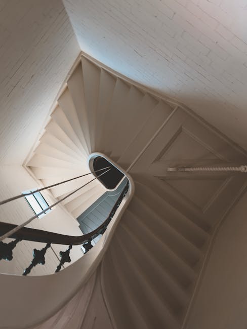 A spiral staircase inside a residential building with white-painted wooden steps and black decorative railing balusters. The staircase curves upward towards a landing near a closed door, with a window on the left side allowing natural light to illuminate the area. The surrounding walls are painted white, and the space appears clean and well-maintained, suitable for home relocation or furniture transport as part of a house removal process. The image was taken from a higher vantage point, providing a clear view of the staircase’s structure and the interior environment, reflecting the interior of a typical property prepared for moving services by Movers Pimlico.
